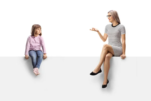 Little girl sitting on a blank panel and listening to a woman talking isolated on white background