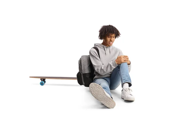 一个拿着滑板的非裔美国人坐在地上 用白色的背景把受伤的膝盖隔离起来 — 图库照片
