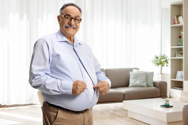 Mature man with big belly buttoning a shirt in a living room at home