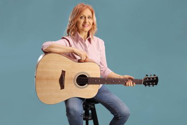 Sandalyede oturan ve elinde akustik gitar tutan bir kadın mavi arka planda izole edilmiş.