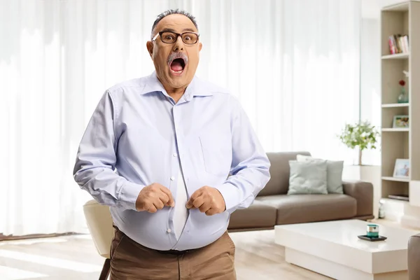 Shocked mature man with a big belly trying to button a shirt in a living room at home