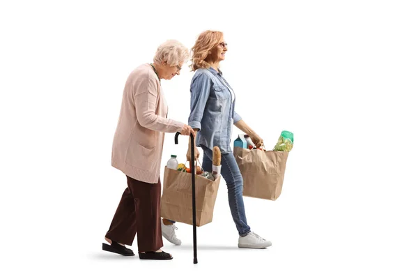 白い背景に隔離された食料品袋を持って杖と若い女性と歩く高齢女性の完全な長さのプロフィールショット — ストック写真