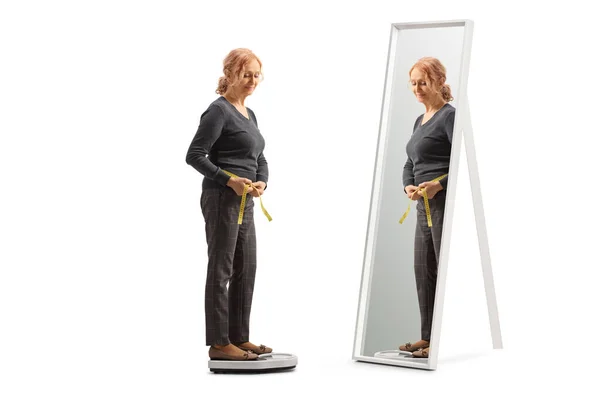 全长拍摄一个成熟的女人用带子测量腰围在一面白色背衬的镜子前面 — 图库照片