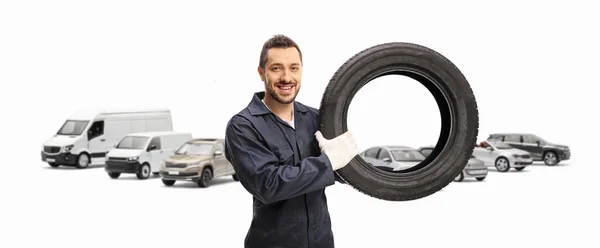 汽车修理工在停放的车辆前拿着车胎 对着白色背景的相机笑着 — 图库照片