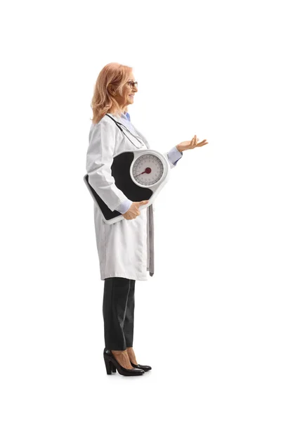 白い背景に隔離された手で体重計とジェスチャーを保持している女性医師の完全な長さプロファイルショット — ストック写真