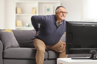 Sırtı ağrıyan yetişkin bir adam evde TV karşısında kanepeden kalkmaya çalışıyor.