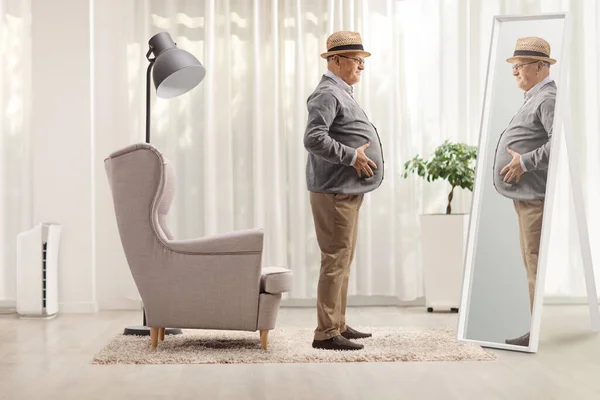 Elderly man with a big belly standing in front of an armchair and looking at a mirror at home