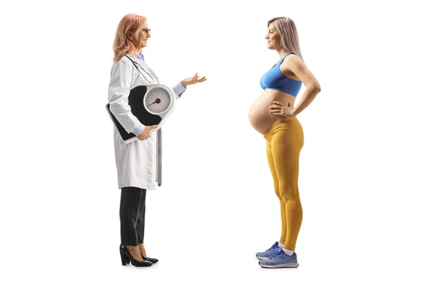 一名女医生举着举重秤 与一名身穿白色背景运动服的孕妇交谈的全身像照片 — 图库照片