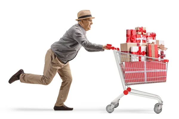白い背景に孤立したプレゼントでいっぱいのショッピングカートで走っている高齢者のフル長さのプロフィールショット — ストック写真