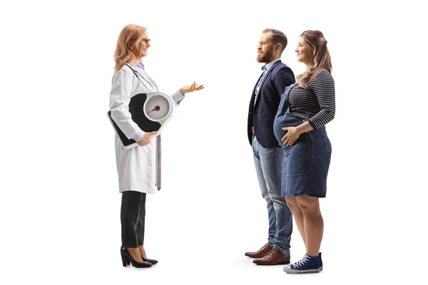 体重計を持っている女性医師の完全な長さのプロフィールショットと白の背景に隔離された妊娠中のカップルと話 — ストック写真