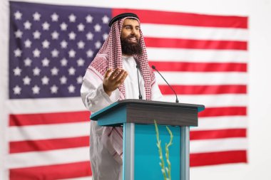 Arap bir adam arka planda ABD bayrağıyla el hareketleriyle bir konuşma yapıyor.