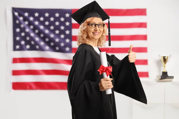 アメリカ国旗の前で修士号を取得し親指を立てる黒の卒業生のガウンに身を包んだ女性 — ストック写真