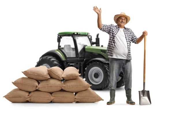 在一辆绿色拖拉机前 一个拿着铁锹和一堆麻袋的成熟农民的全长肖像 微笑着 在白色背景下挥动着孤零零的手 — 图库照片