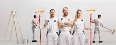 Ev dekoratörleri ayakta kameraya bakıyor ve diğeri de duvarı boyuyor.