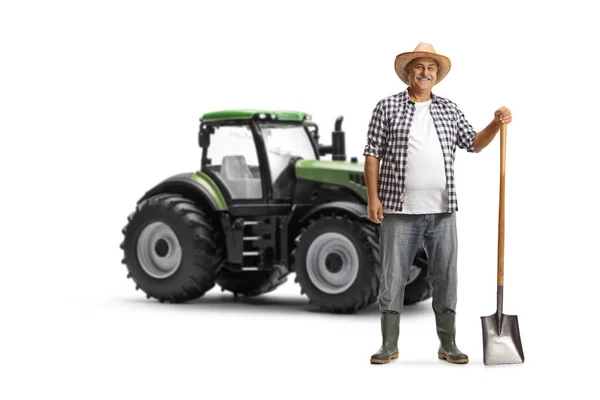 成熟的农民 拿着铲子站在一辆绿色拖拉机旁边 在白色的背斜上微笑着 — 图库照片