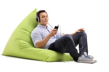 Genç adam koltukta oturmuş, beyaz arka planda izole edilmiş kulaklıklar ve akıllı telefonlarla müzik dinliyor.