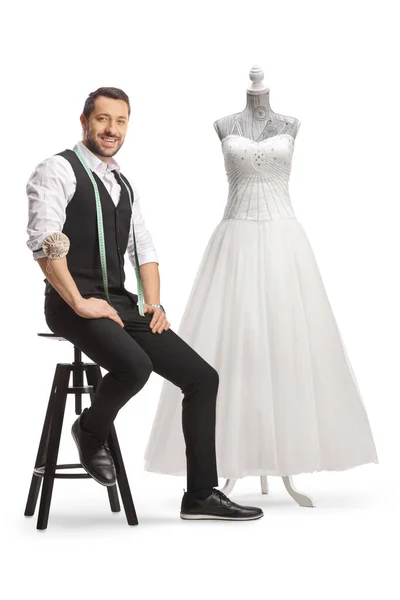年轻的裁缝 带着测量带 对着相机笑着 坐在白色背景的婚纱旁边 — 图库照片