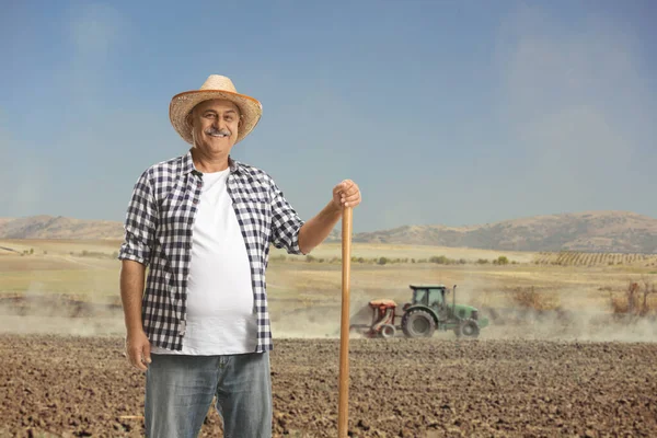 一个成熟的农民 拿着铲子在地里摆姿势 后面坐着一辆拖拉机 — 图库照片