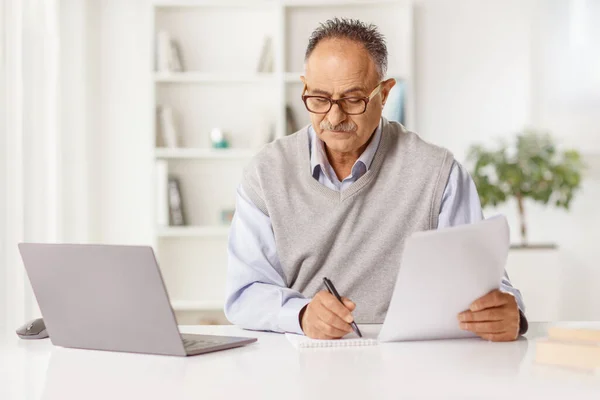 成熟した男は自宅でノートパソコンの前に座って 紙の文書を書く — ストック写真