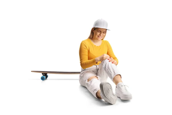 若いです女性とともにスケートボード座っています床と保持彼女の負傷膝隔離された上の白い背景 — ストック写真