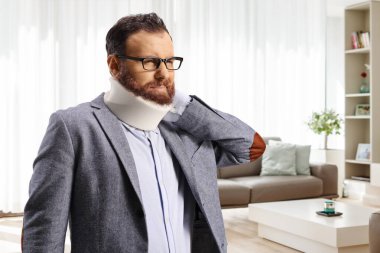 Sakallı, boyun tasmalı, ağrılı boynunu evdeki bir oturma odasında tutan adam.