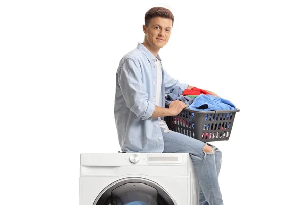 一个拿着洗衣篮的年轻人坐在一台白色背景的洗衣机上 — 图库照片