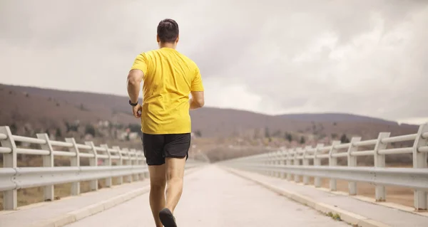 在阴天 一个身穿黄色T恤衫的男子在桥上慢跑时的后听镜头 — 图库照片