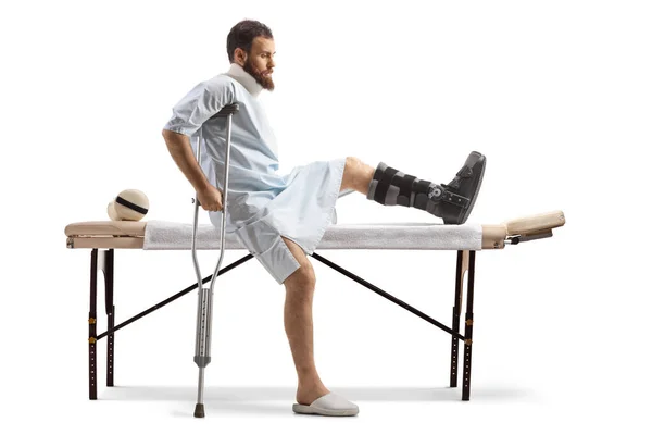 白い背景に隔離された壊れた足と首の襟と医療テーブルに座っている病院のガウンの男 — ストック写真