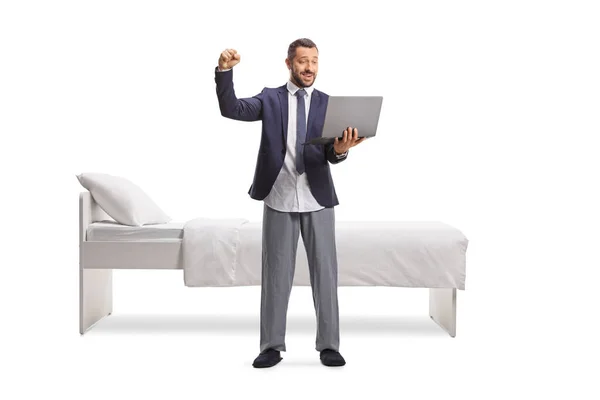 穿着西服 穿着底睡衣的男人 用笔记本电脑 在被白色背景隔开的床前为自己的幸福做手势 — 图库照片