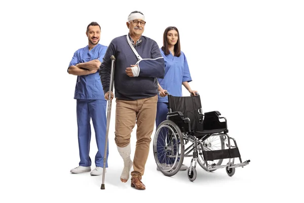 車椅子の青い制服を着た医療従事者と白い背景に隔離された松葉杖と腕のスリングを持つ成熟した男性患者 — ストック写真