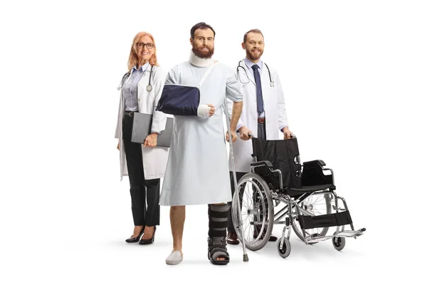 白い背景に隔離された医師のチームと一緒に立っている壊れた足と腕を持つ病院のガウンの髭の男 — ストック写真