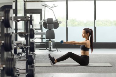 Paspasa oturmuş spor salonunda dambıllarla egzersiz yapan genç bir kadın.
