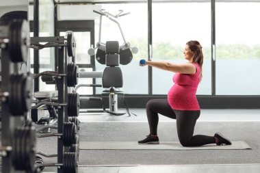 Hamile bir kadının spor salonundaki paspasın üstünde dambıllarıyla egzersiz yaptığı uzun bir profil çekimi.