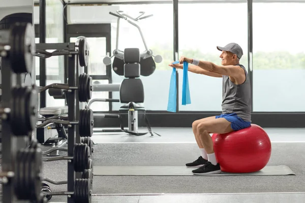 身穿运动服的老年男子坐在一个健身球上 带着一个橡皮筋在体育馆里锻炼 — 图库照片