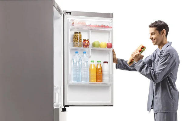 パジャマ姿の男がサンドイッチを食べ白い背景に隔離された冷蔵庫を開ける — ストック写真