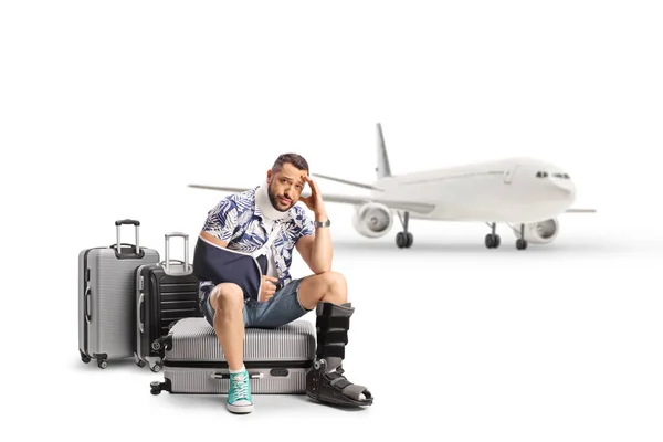 白い背景に隔離された飛行機の前に負傷した腕と脚がスーツケースに座っている悲しい男性観光客 — ストック写真