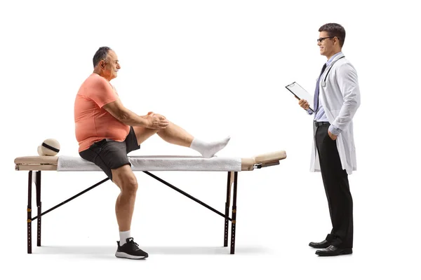 マッサージベッドの上に座って 白い背景に隔離された医師と話す痛みを伴う膝を持つ成熟した屈強な男 — ストック写真