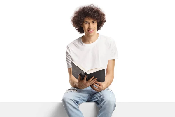 Młody Człowiek Kręconymi Włosami Siedzący Panelu Trzymający Książkę Białym Tle — Zdjęcie stockowe