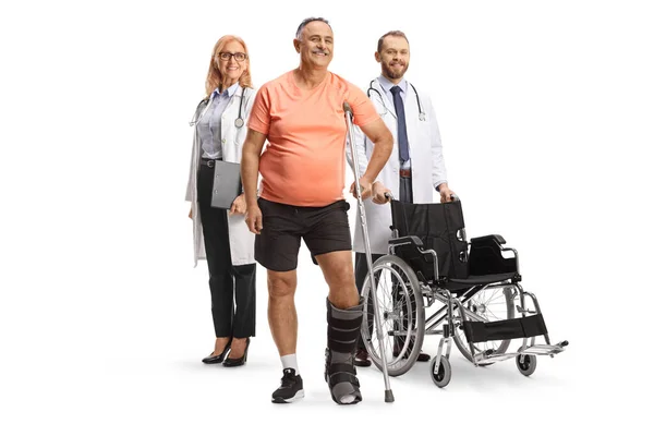 車椅子の医者と整形外科のブーツと白い背景に隔離された松葉杖を持つ成熟した男性患者 — ストック写真