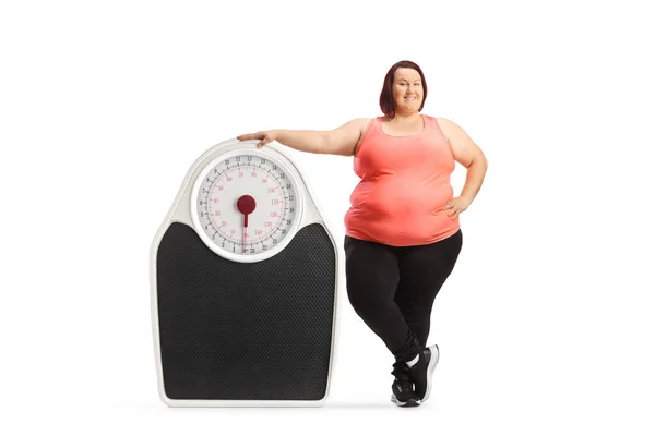 白い背景に隔離された大きな体重計の横に立ってスポーツウェアで太りすぎの女性 — ストック写真