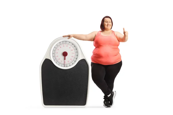 スポーツウェア姿の太りすぎの女性が大きな体重計の横に立っているとジェスチャー親指アップ上の白い背景に隔離 — ストック写真