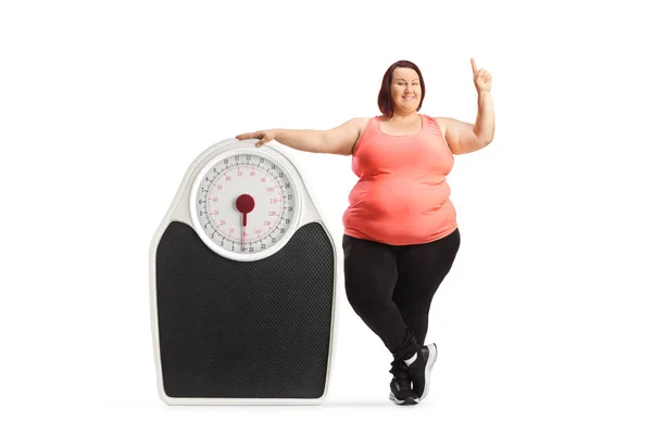 スポーツウェア姿の太りすぎの女性が大きな体重計の横に立って 白い背景に隔離された上を指して — ストック写真