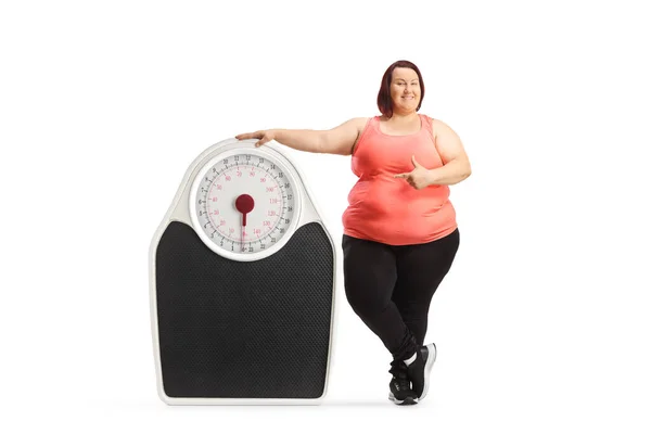 スポーツウェア姿の太りすぎの女性は 大きな体重計の横に立って 白い背景に隔離された指す — ストック写真