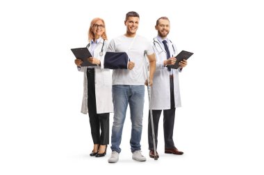 Koltuk değneği ve kol askısı olan genç bir adam beyaz arka planda izole edilmiş bir doktor ekibinin önünde duruyor.