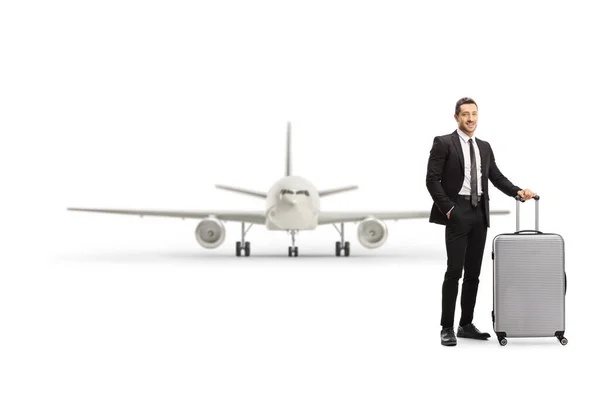 白い背景に隔離された航空機の前でスーツケースを身に着けているスーツケースに身を包んだ若い男の完全な長さの肖像 — ストック写真