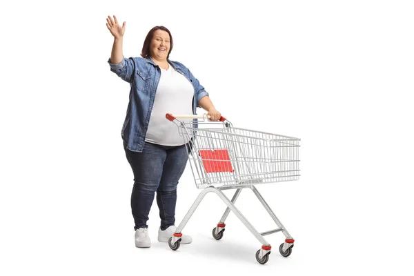 ショッピングカートの笑顔で太りすぎの女性の完全な長さショットと白の背景に隔離された手を振る — ストック写真