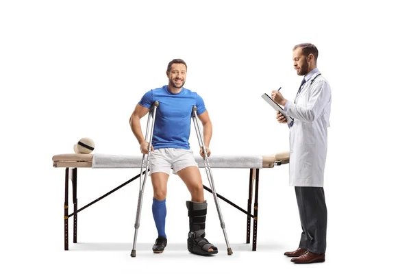 白い背景に隔離された文書を書く医者と理学療法ベッドの上に座って松葉杖を持つサッカー選手 — ストック写真