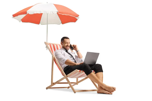 ビジネスマンは 傘の下でデッキチェアに座って 白い背景に隔離されたラップトップコンピュータとスマートフォンを使用しています — ストック写真