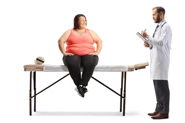 白い背景に隔離された理学療法ベッドに座っている太りすぎの女性に処方書を書く医師 — ストック写真