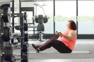 Kilolu bir kadın spor salonunda direniş bandıyla egzersiz yapıyor.
